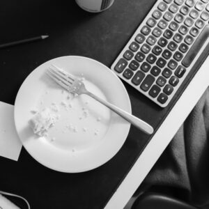 Topshot eines Schreibtischs. Eine Tastatur steht im Anschnitt neben einem weißen Teller. Auf dem Teller liegt ein Kuchen und eine Gabel. Um Diesen Teller liegen Stifte und Zettel.